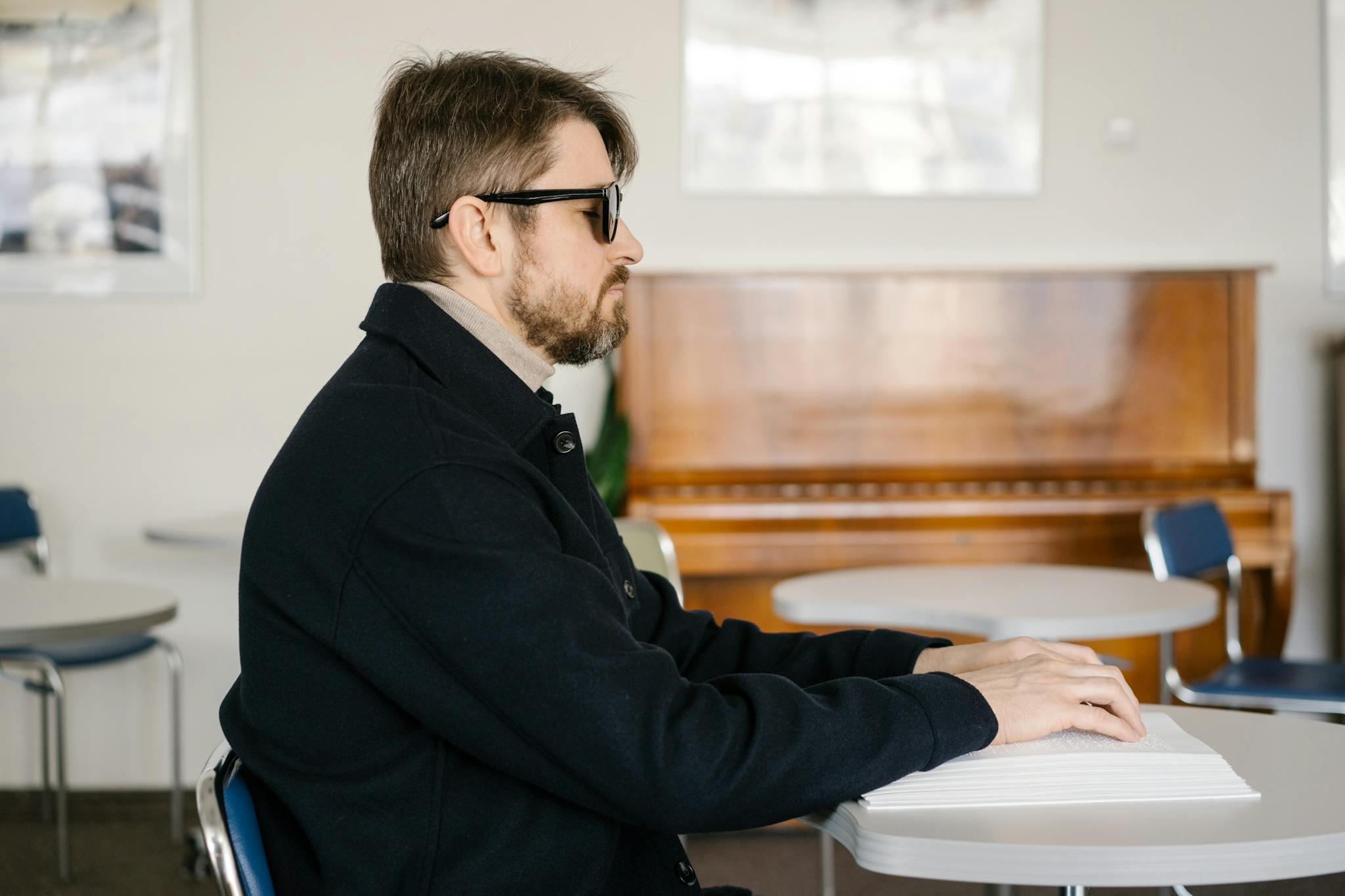 A visually impaired man reading Braille at a table in a warmly lit room with a piano in the background.