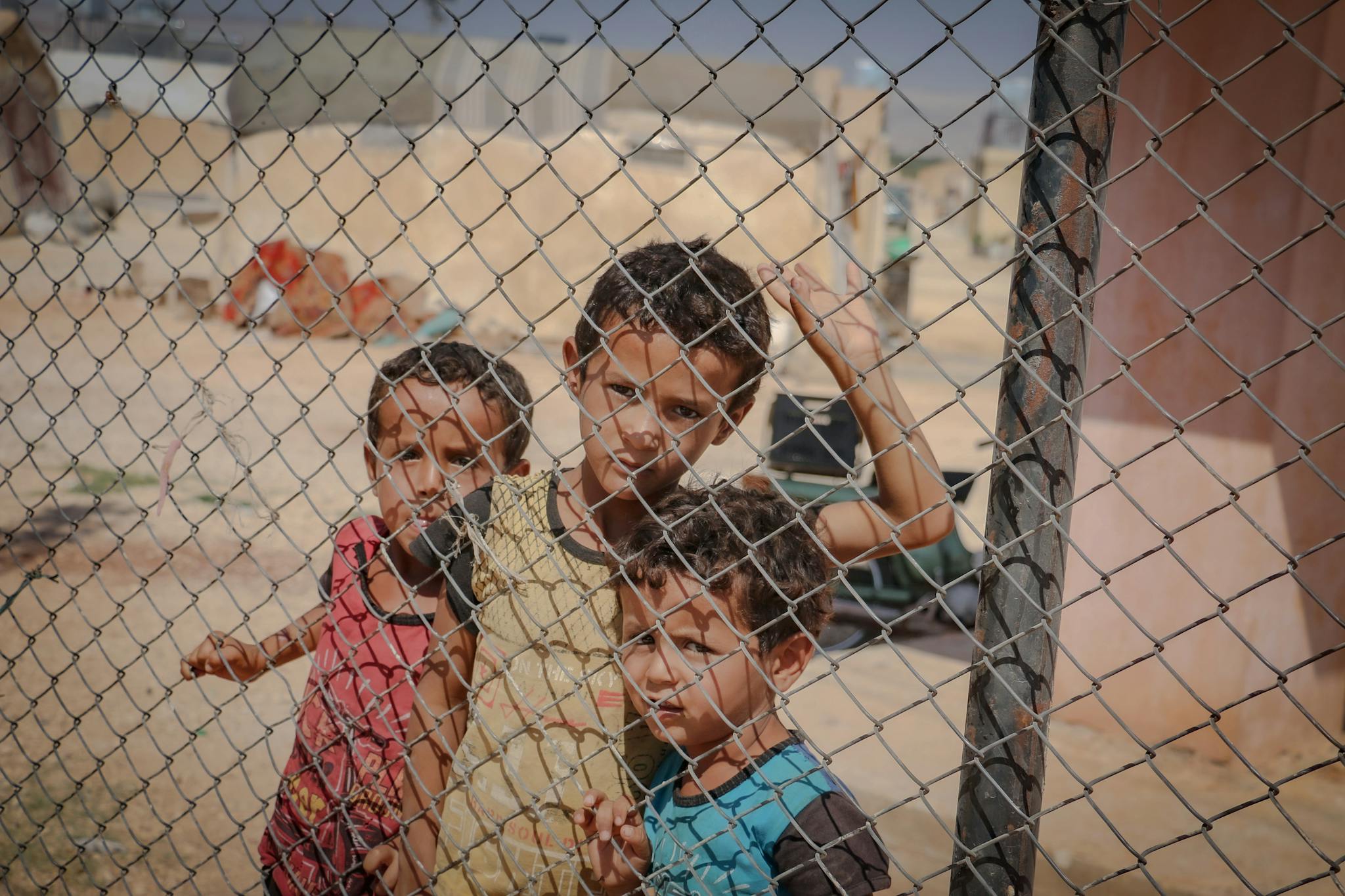 Heartfelt image of children behind a fence in a Syrian refugee camp, under a sunny sky.